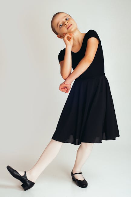 Full body calm girl in black dress for dancing looking at camera while touching face and stand foot on heel against white background
